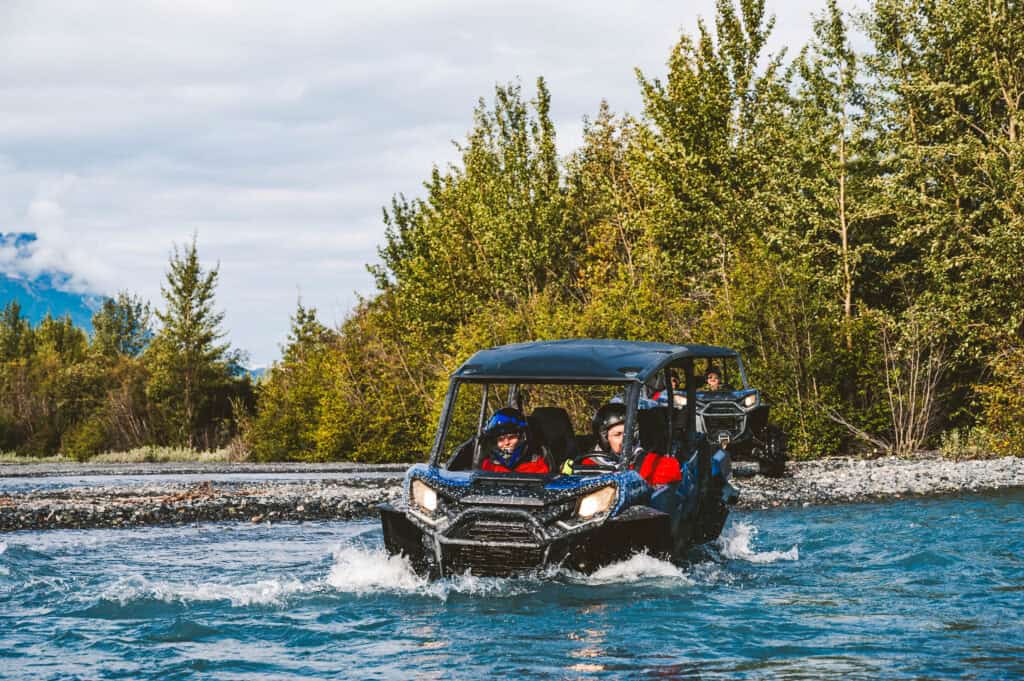 A CanAm Side-by-Side Crossing an Alaskan River as driver and passenger smile.