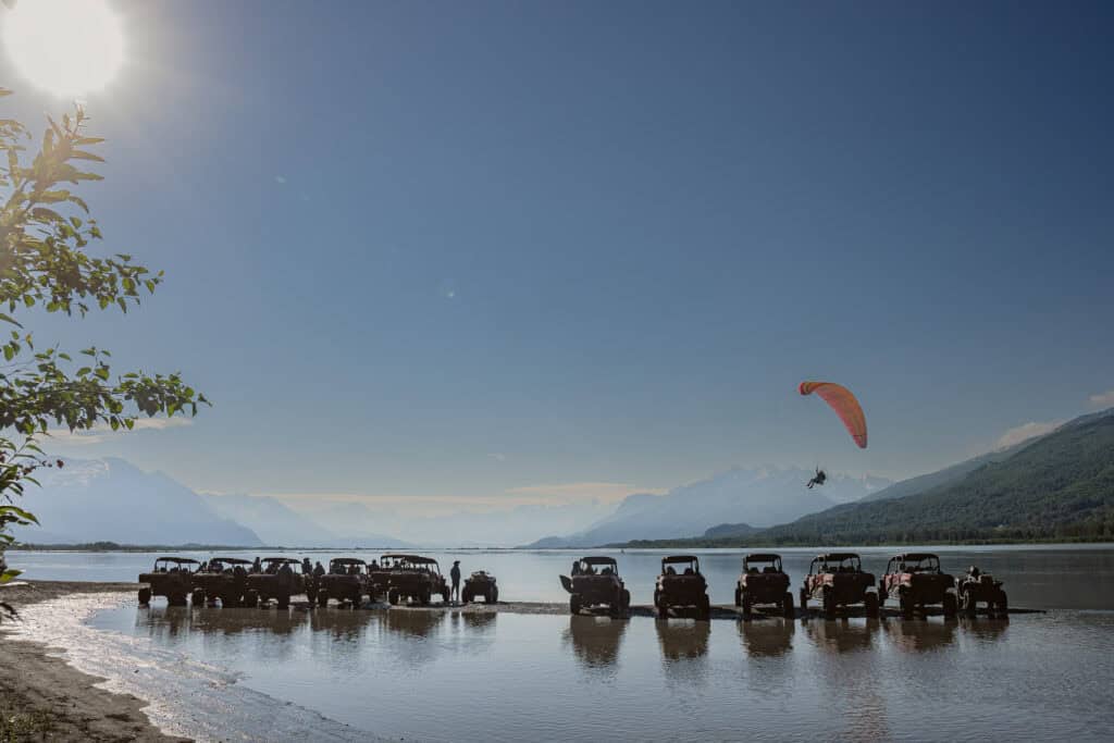 atvs lined up in the knik river while a paraglider flies overhead